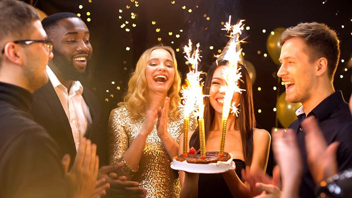 Friends celebrating a special occasion at Asha's with a birthday cake adorned with sparklers, featuring people laughing and clapping in festive attire against sparkling lights background