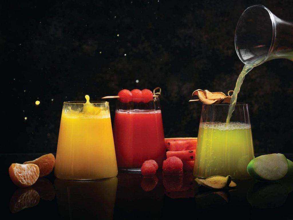 Fresh cold-pressed juices at Asha's Restaurant - orange, watermelon, and green apple juices garnished with fresh fruits on dark background